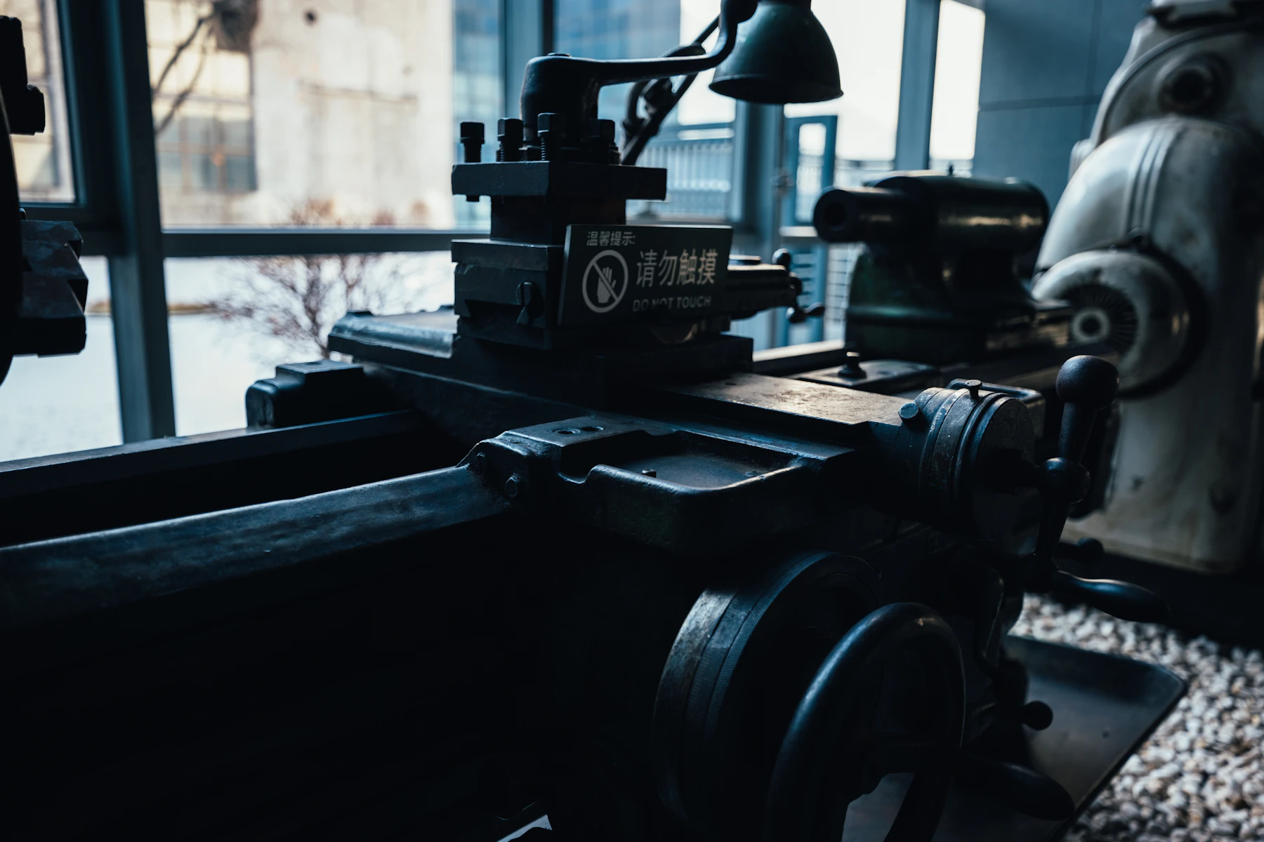 A metal lathe and its precision controls beside tall workshop windows